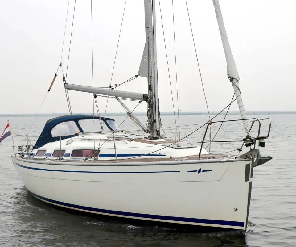 Een witte zeilboot met blauwe strepen en de naam "Diva" op de romp, vaart op rustig water onder een grijze lucht.