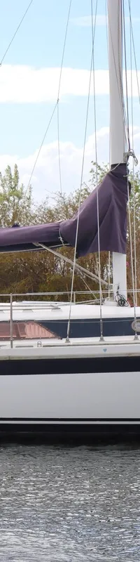 Een witte zeilboot met donkerblauwe streep en een opgerold paars zeil vaart op het water, met een persoon aan het roer.