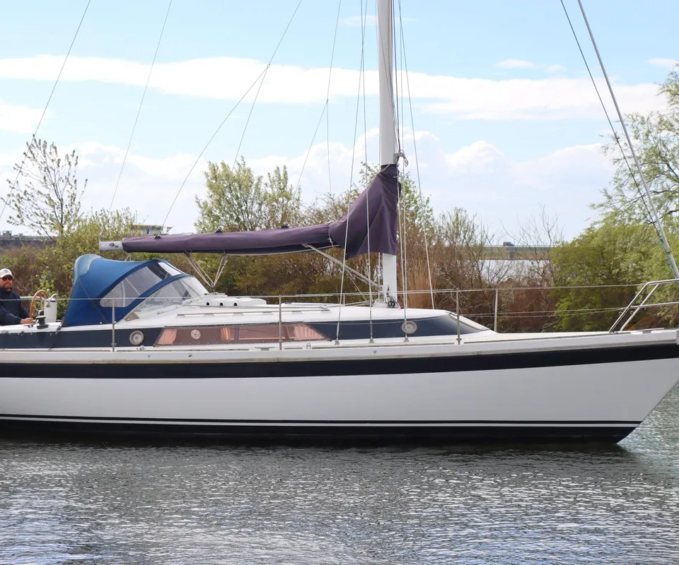 Een witte zeilboot met donkerblauwe streep en een opgerold paars zeil vaart op het water, met een persoon aan het roer.