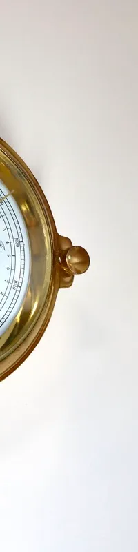 Een gouden barometer en een gouden lampje zijn gemonteerd op een witte muur, met een houten plank erboven.