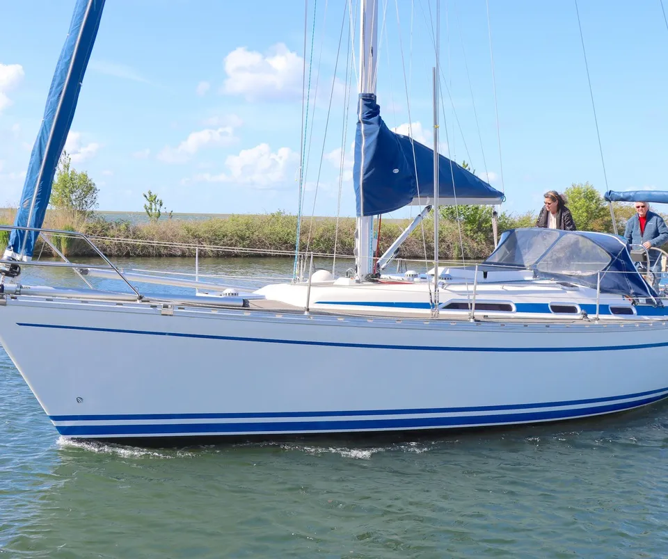 Een witte zeilboot met blauwe strepen vaart op het water, met twee mensen aan boord en een blauw zeil. — Bavaria 38