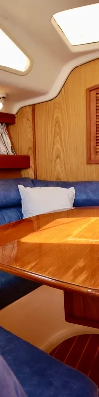 De foto toont het interieur van een boot met een houten tafel, blauwe banken, houten wanden en een doorgang naar een…