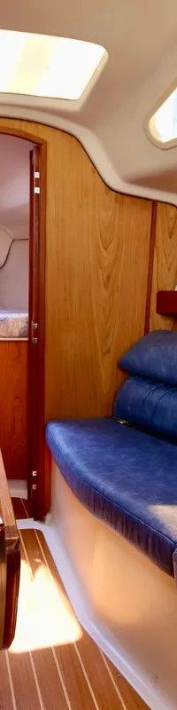 De foto toont het interieur van een boot met een houten tafel, blauwe banken en een doorgang naar een slaapcabine.