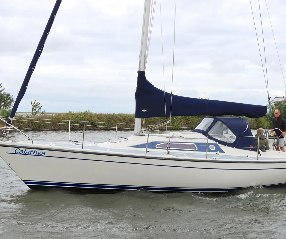 Een witte zeilboot met blauwe accenten en de naam "Calathea" vaart op water, met twee personen aan boord.