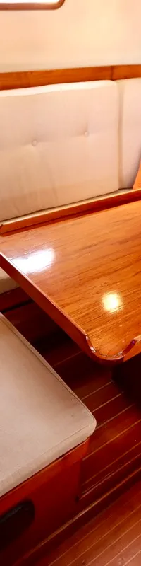 De foto toont een kajuitinterieur met een U-vormige bank met lichte kussens, een houten tafel en een blauwe lamp.