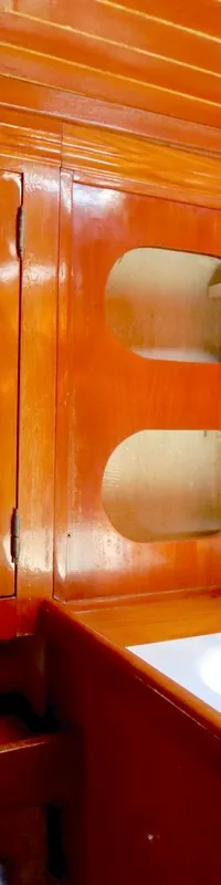 Een houten interieur met een wasbak, twee kranen, een ronde spiegel en kasten met jaloezieën en open vakken.