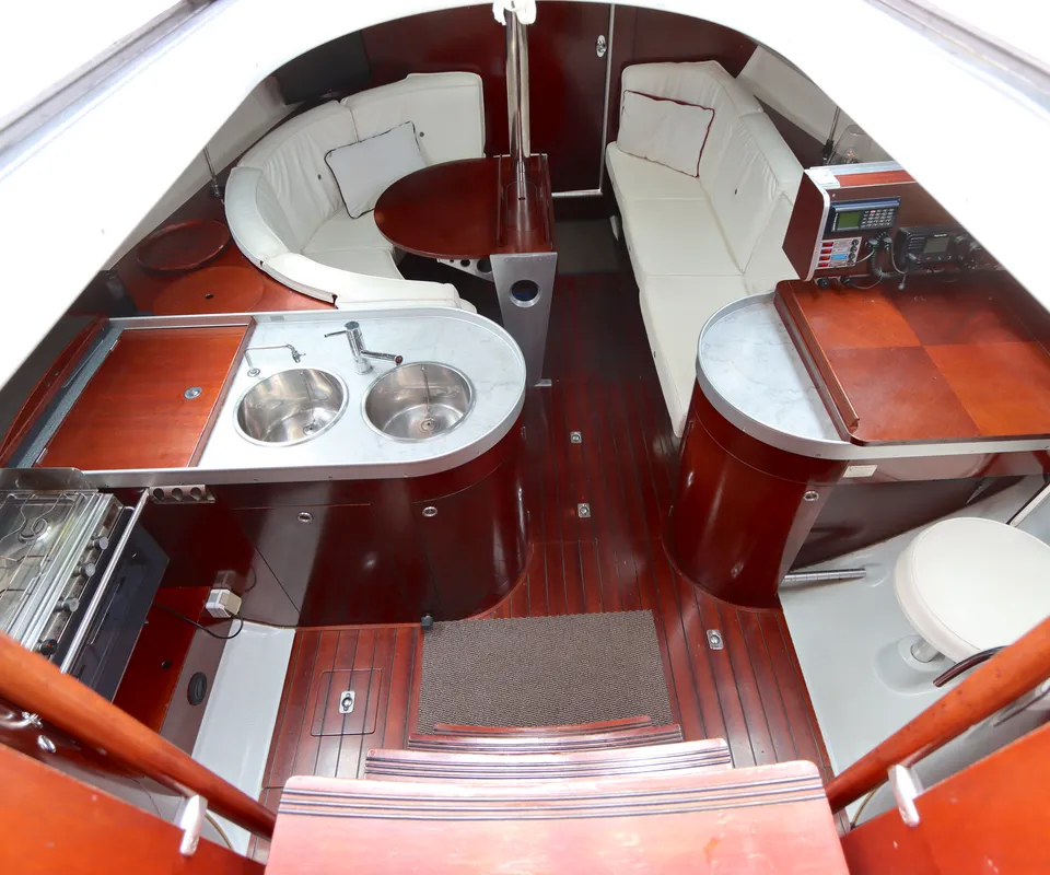 De foto toont het interieur van een boot met een kombuis, zithoek en navigatiehoek, alles afgewerkt met donker hout en…
