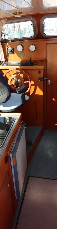 De kajuit toont een houten interieur met stuurstand, kombuis met twee kookplaten, een zithoek met blauwe kussens en een…