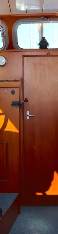 De kajuit toont een houten interieur met een stuurstand, een blauwe stoel, een kombuis met kookplaat en spoelbak, en een…