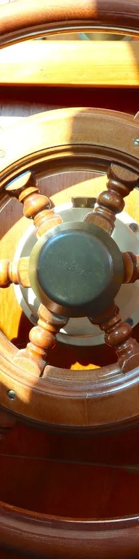 Een houten scheepsstuurwiel met acht spaken en handvatten is te zien in een houten interieur, verlicht door zonlicht.