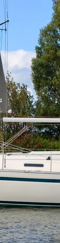 Een crèmekleurige zeilboot met groene strepen en een groene buiskap vaart op het water, met de naam "Godewind" op de boeg…