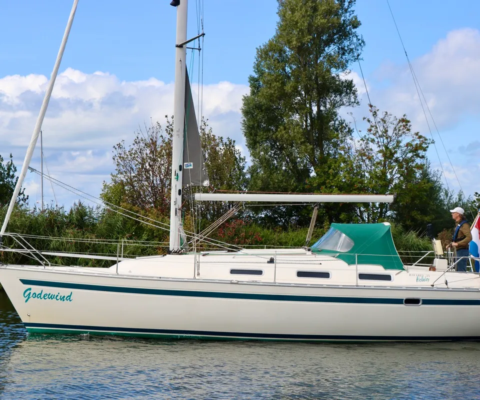 Een crèmekleurige zeilboot met groene strepen en een groene buiskap vaart op het water, met de naam "Godewind" op de boeg…