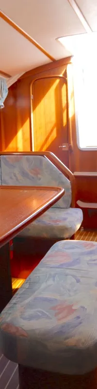 Een houten tafel met banken met stoffen bekleding staat in de salon, met een kombuis en ramen zichtbaar.