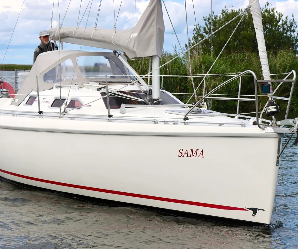 Een witte zeilboot met de naam "SAMA" en een rode streep op de romp vaart op het water, met een persoon aan boord. — Etap 30i
