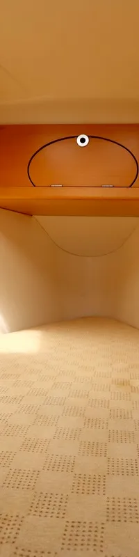 De kajuit toont een V-vormig bed met een lichtgekleurd matras, omringd door houten kasten en twee netten aan de wanden.