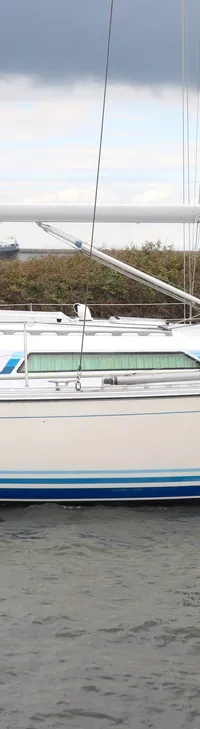 Een witte zeilboot met blauwe strepen op de romp en de naam "Hunt" vaart op het water, met een persoon aan het roer.