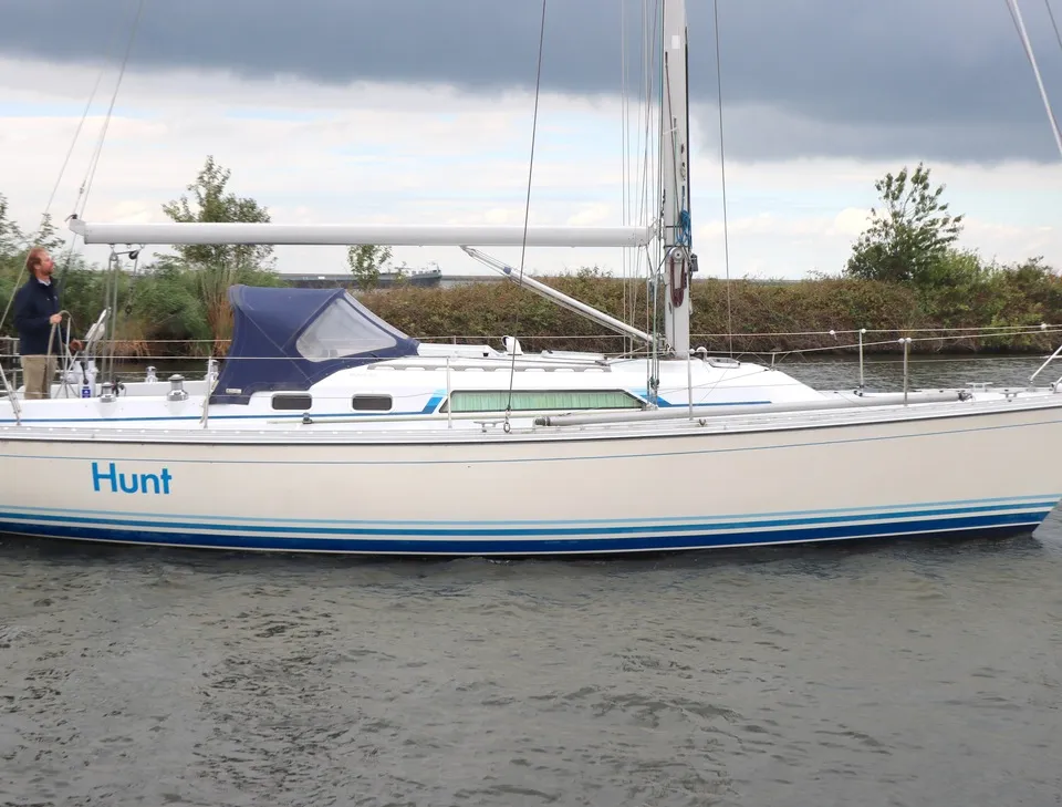 Een witte zeilboot met blauwe strepen op de romp en de naam "Hunt" vaart op het water, met een persoon aan het roer.
