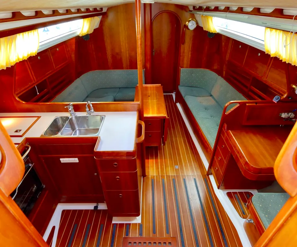 De foto toont een houten bootinterieur met een kombuis, twee banken met groene kussens en een houten vloer.
