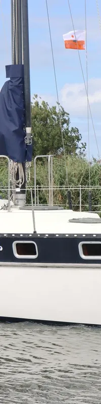 Een witte zeilboot met donkerblauwe band en de naam "PHANTOM" vaart op het water, met een persoon in de kuip en vlaggen.