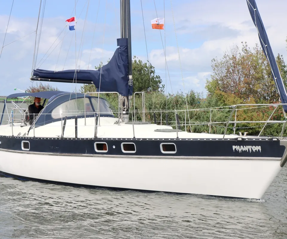 Een witte zeilboot met donkerblauwe band en de naam "PHANTOM" vaart op het water, met een persoon in de kuip en vlaggen.
