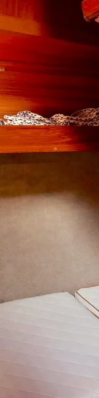 De kajuit toont houten betimmering, een raam met gordijn, een rode brandblusser, witte matrassen en een oranje kist.