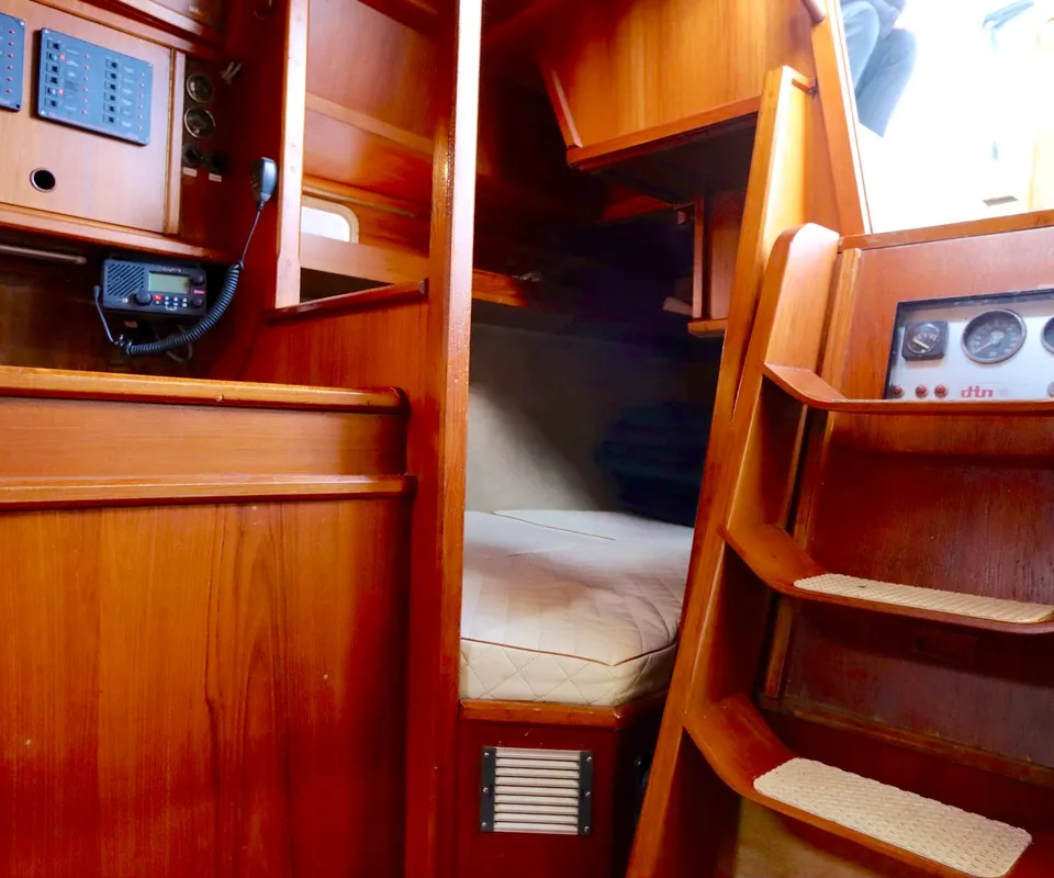 De foto toont een houten bootinterieur met een bed, een marifoon, schakelpanelen, meters en een trap.