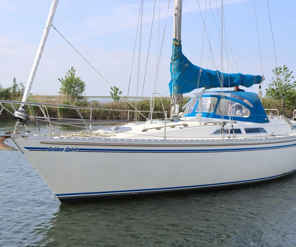 Een witte zeilboot met blauwe strepen en de naam "Golden Spirit" op de romp ligt in het water, met een blauwe sprayhood en…