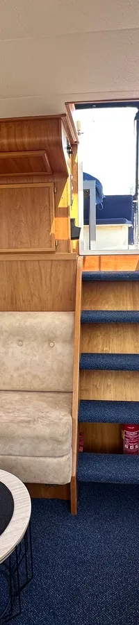 Een interieur van een boot met een beige L-vormige bank, een blauw tapijt, houten wanden en een trap naar boven.