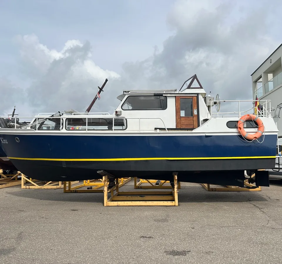 Een blauw-witte motorboot met de naam "Kira" op de boeg staat op gele bokken onder een bewolkte hemel. — Altena GSAK