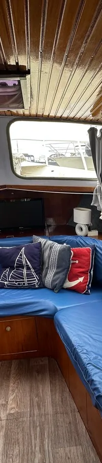 De kajuit heeft een houten plafond, blauwe U-vormige zitbanken, een houten vloer en ramen met grijze gordijnen. — Altena GSAK