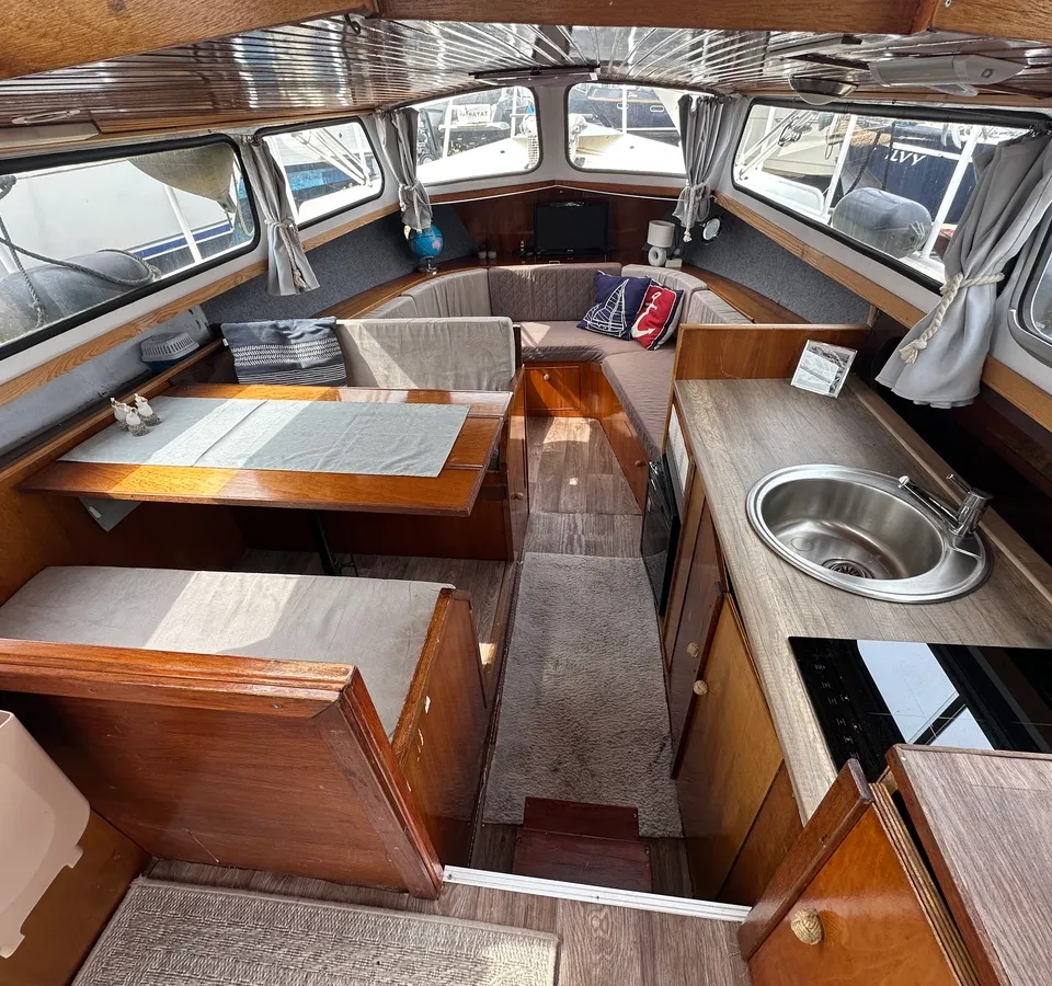 De foto toont het interieur van een boot met houten afwerking, een zithoek met tafel, een kombuis met spoelbak en…