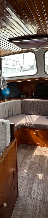 De kajuit toont een houten plafond, ramen met gordijnen, een U-vormige zithoek met grijze kussens en een houten tafel.