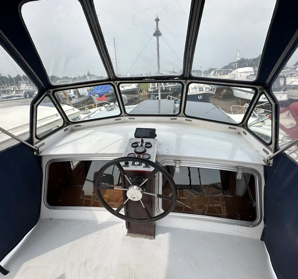 De foto toont het stuurgedeelte van een boot met een stuurwiel, instrumentenpaneel en een voorruit met meerdere ramen.