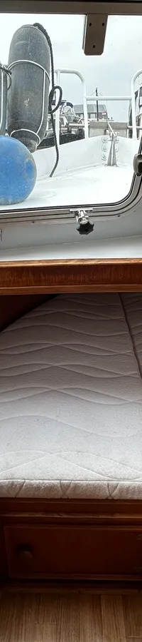 Een interieurfoto van een bootkajuit met een wit matras, houten afwerking en ramen die uitkijken op andere boten.