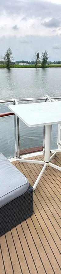 Een houten dek met twee grijze zitmeubels en een witte tafel aan boord van een boot, met water en landschap op de…