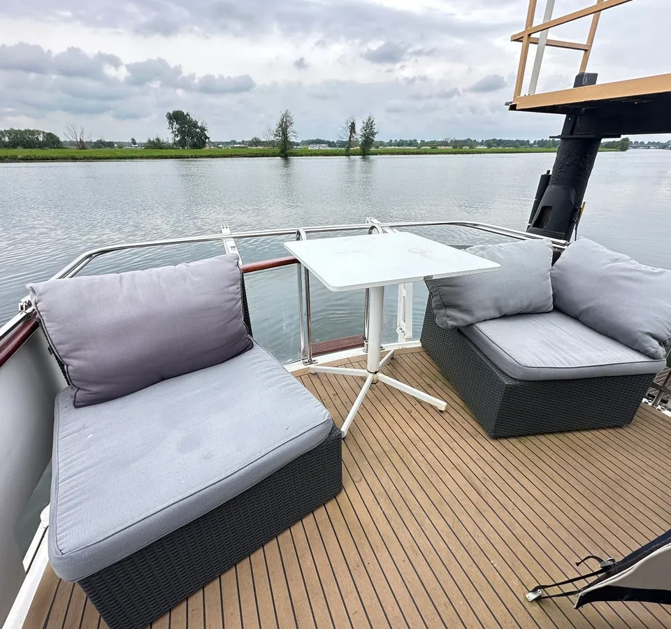Een houten dek met twee grijze zitmeubels en een witte tafel aan boord van een boot, met water en landschap op de…