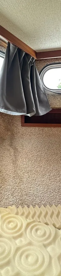 Een interieur van een boot met een bed met een crèmekleurig matras, houten wanden en ramen met grijze gordijnen.