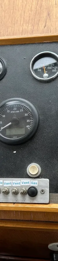 Een houten dashboard met een zwart paneel met diverse meters, schakelaars en een bedieningspaneel voor de boegschroef.