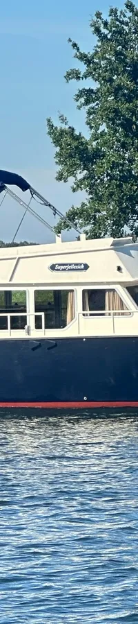 Een donkerblauwe motorboot met een crèmekleurige opbouw en een rood onderwaterschip vaart op een waterweg.