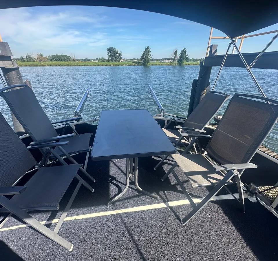 Een dek met een tafel en vier stoelen, met uitzicht op water en een groene oever.