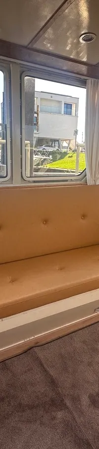 Een interieur van een boot met een bruine bank, een stuurwiel en ramen die uitzicht bieden op water en andere boten.