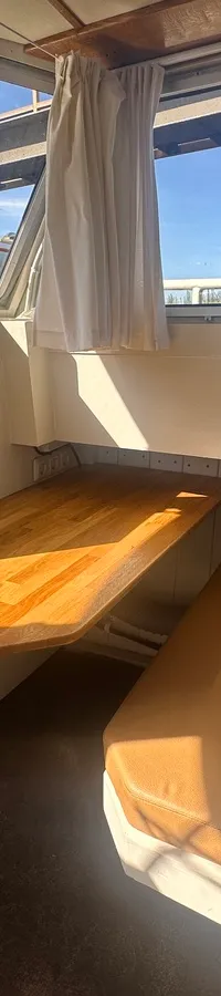 Een interieur van een boot met een houten tafel, een bank met bruine kussens en ramen met witte gordijnen.