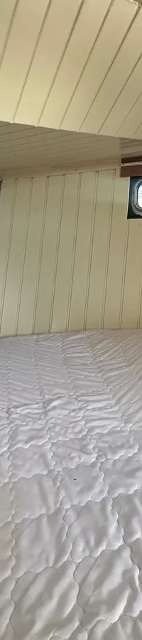 Een kajuit met een groot wit matras, twee kleine ramen met gordijnen en een wit houten plafond en wanden. — Oomen kruiser fly