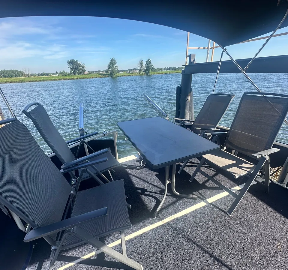 Een dek met vier donkere stoelen en een donkere tafel, met uitzicht op water en een groene oever.
