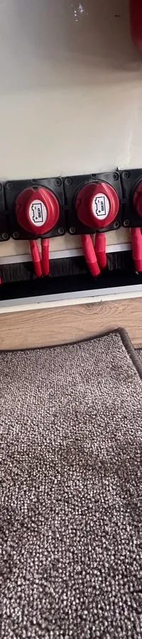 Zes rode hoofdschakelaars met rode kabels zijn gemonteerd op een witte wand boven een grijze vloerbedekking.