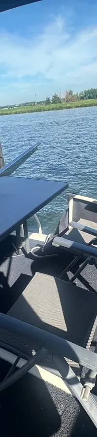 Een dek met een tafel en vier stoelen onder een zonnescherm, met water en een landschap op de achtergrond.