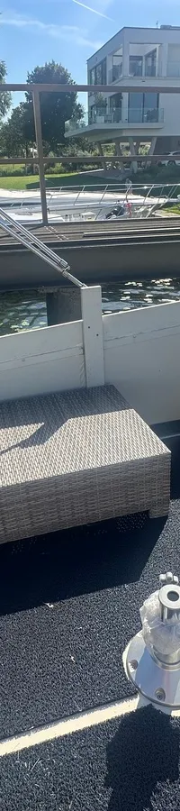 Een achterdek van een boot met een rieten bank, twee open luiken en een lier, met water en gebouwen op de achtergrond.