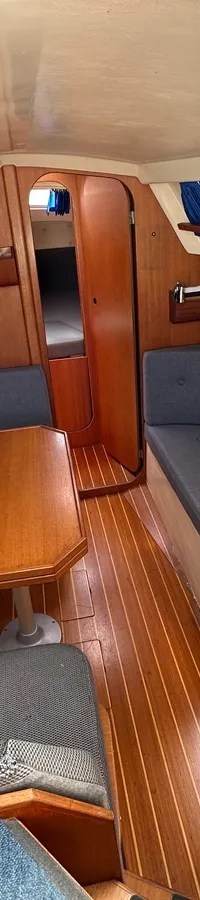 De foto toont het interieur van een boot met houten vloer en wanden, grijze banken, een tafel en een kombuis met fornuis en…