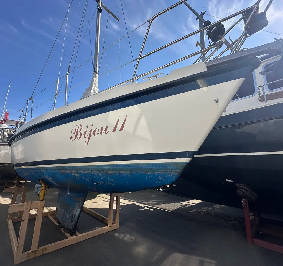 Een witte zeilboot met de naam "Bijou II" in rode letters staat op houten bokken naast een donkere boot. — Compromis 777