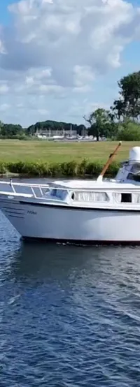Een witte motorboot vaart op blauw water met een groene oever en bomen op de achtergrond onder een bewolkte hemel.